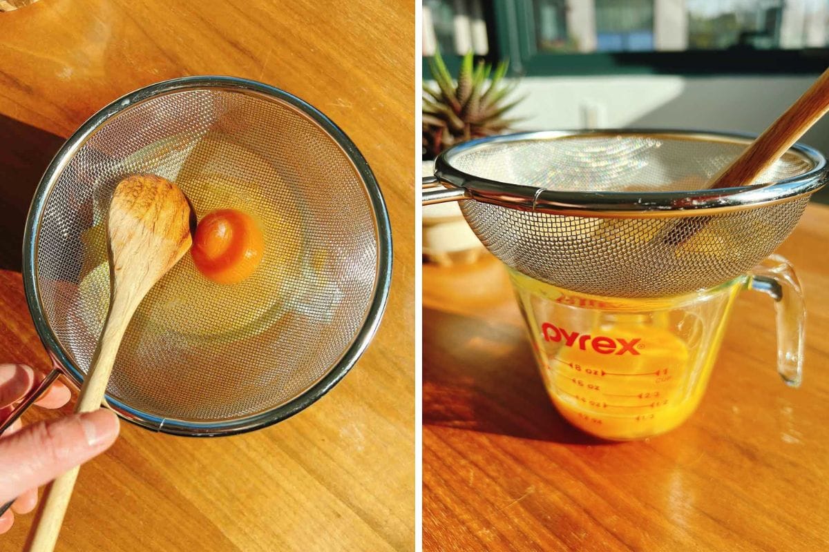 A hand uses a wooden spoon to press an egg through a metal strainer for egg drop noodle soup; beside it, yolks and whites are strained into a Pyrex measuring cup on a sunlit wooden table.