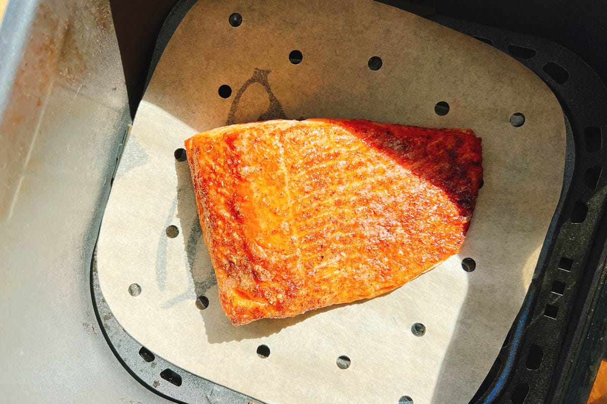 An air fried fillet of trout sits on parchment paper inside an air fryer basket, with light highlighting its crispy, golden-brown surface—demonstrating how perfectly you can prepare air fryer trout or salmon for a delicious meal.
