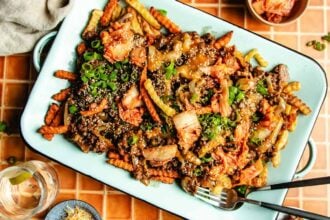 A platter of Bulgogi Fries—crinkle-cut fries topped with beef mince, kimchi, green onions, and sesame seeds—sits on a tiled table, with a fork and a glass of water nearby.