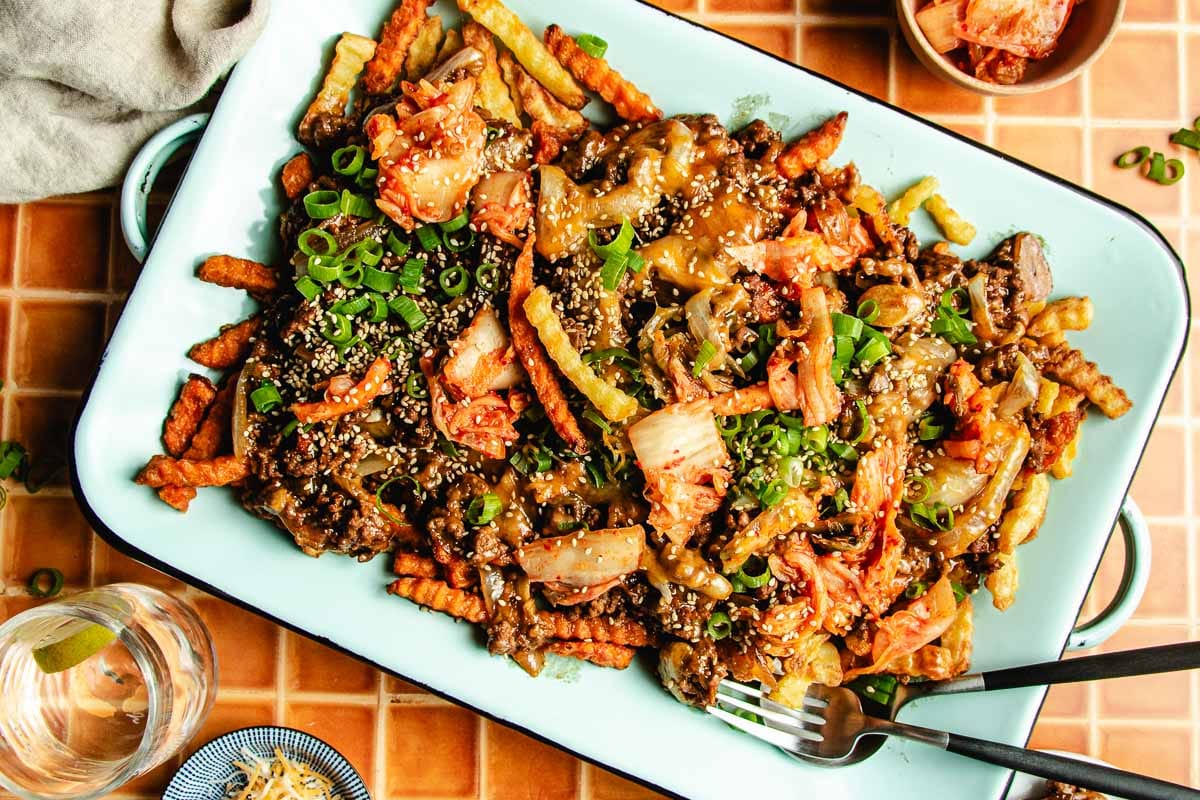 A platter of Bulgogi Fries—crinkle-cut fries topped with beef mince, kimchi, green onions, and sesame seeds—sits on a tiled table, with a fork and a glass of water nearby.
