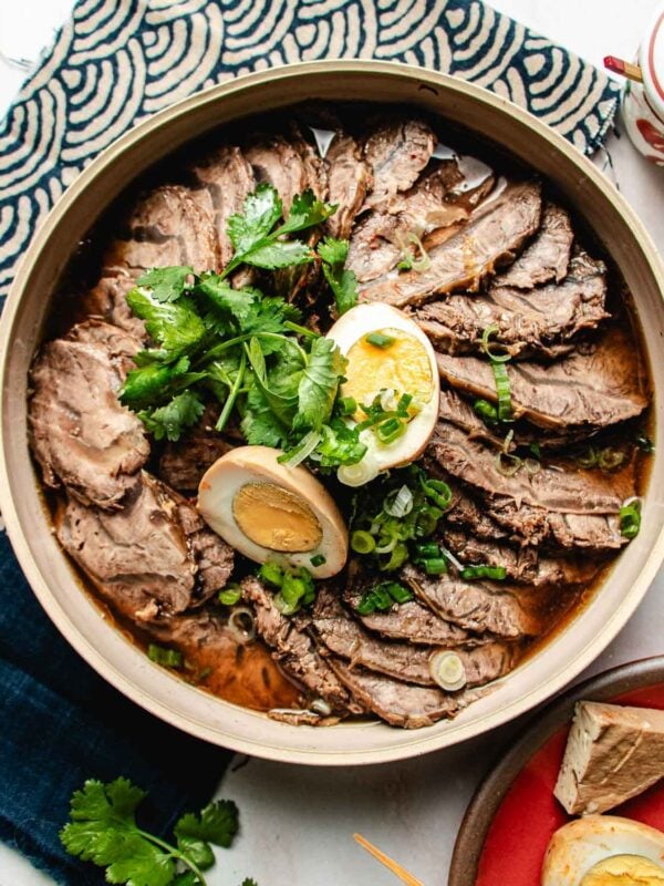 A bowl of Taiwanese Braised Beef Shank in braising sauce, topped with fresh cilantro, green onions, and halved boiled eggs, is placed on a white surface with a patterned cloth nearby.