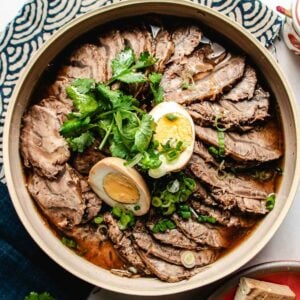 Sliced Taiwanese Braised Beef Shank arranged in a plate with braising sauce broth, topped with fresh cilantro, chopped green onions, and halved soy-marinated eggs, set on a patterned cloth.