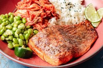 A plate with air fryer trout, steamed white rice topped with black sesame seeds, edamame, kimchi, and lime wedges. The food is arranged neatly and looks fresh and appetizing.