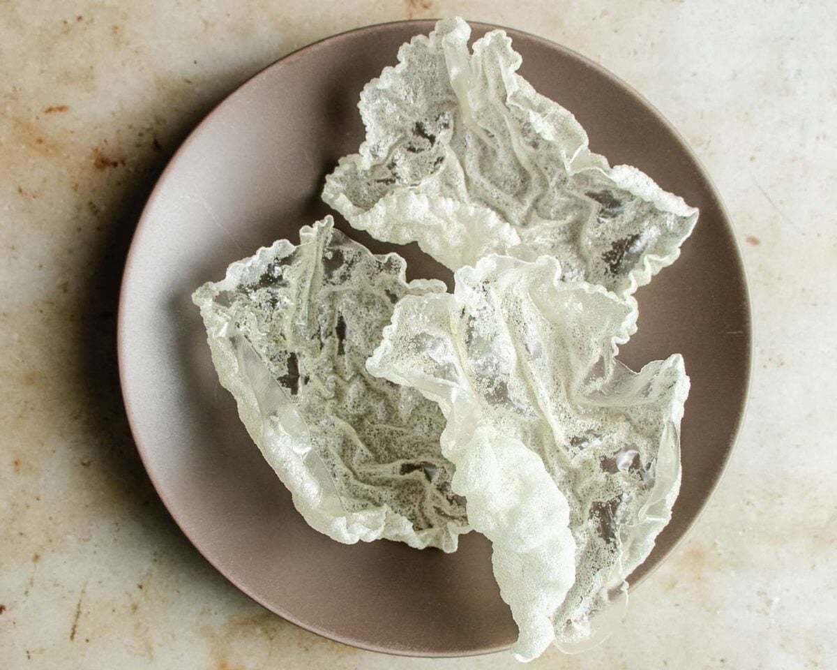 Three pieces of crispy, white puffed rice paper are arranged on a round beige plate, placed on a light-colored, textured surface—an inventive twist with real crunch to add to spring roll in a bowl.