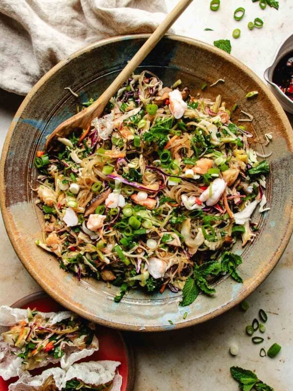 A large bowl of spring roll in a bowl with shrimp, shredded vegetables, fresh herbs, and chopped green onions, mixed with a wooden spoon. A crispy spring roll in a bowl sits nearby on a light surface beside a small plate and cloth with a dressing on the side.