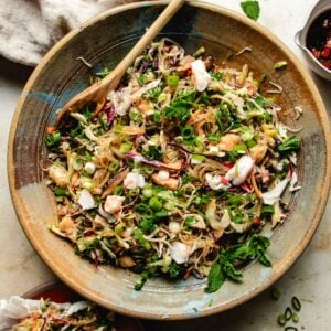 A large bowl filled with a vibrant spring roll in a bowl —featuring shrimp, chopped vegetables, greens, fresh herbs, and green onions, crispy rice paper puffs, with a wooden spoon resting inside.