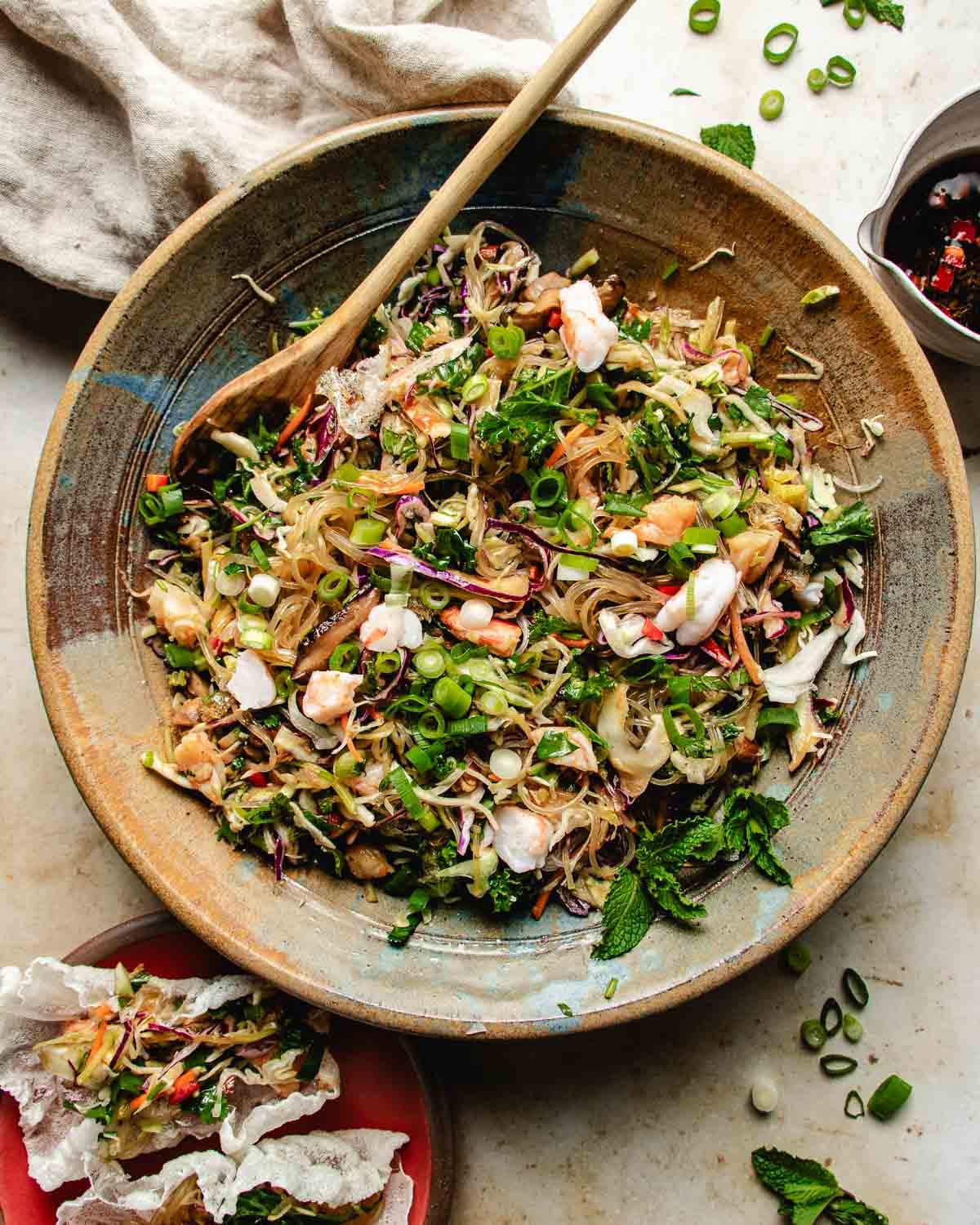 A large bowl of spring roll in a bowl with shrimp, shredded vegetables, fresh herbs, and chopped green onions, mixed with a wooden spoon. A crispy spring roll in a bowl sits nearby on a light surface beside a small plate and cloth with a dressing on the side.