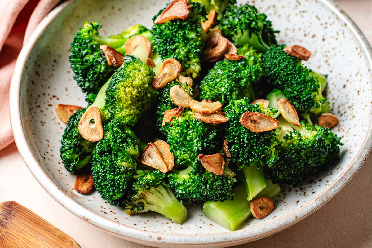 A close-up of a speckled bowl filled with stir fried broccoli florets topped with golden-brown slices of fried garlic chips, reminiscent of Din Tai Fung Stir Fried Broccoli.