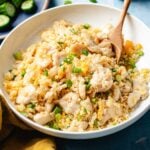 A bowl of Din Tai Fung Chicken Fried Rice with green onions, served with a wooden spoon. The dish features tender chicken, egg, and rice, garnished with chopped green onions. Sliced cucumbers are visible in the background.