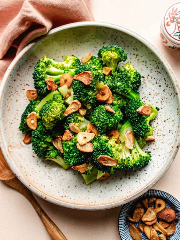 A ceramic plate with Stir fried broccoli florets, inspired by Din Tai Fung Stir Fried Broccoli, topped with golden-brown garlic slices. A wooden spoon, a pink napkin, and a small dish with extra garlic slices are nearby.