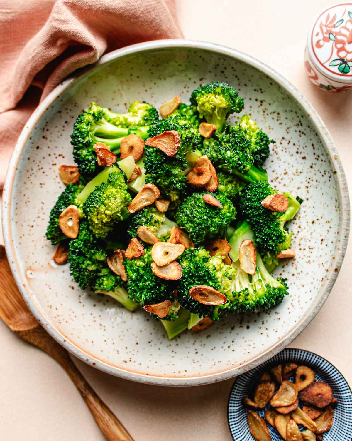 A ceramic plate with Stir fried broccoli florets, inspired by Din Tai Fung Stir Fried Broccoli, topped with golden-brown garlic slices. A wooden spoon, a pink napkin, and a small dish with extra garlic slices are nearby.