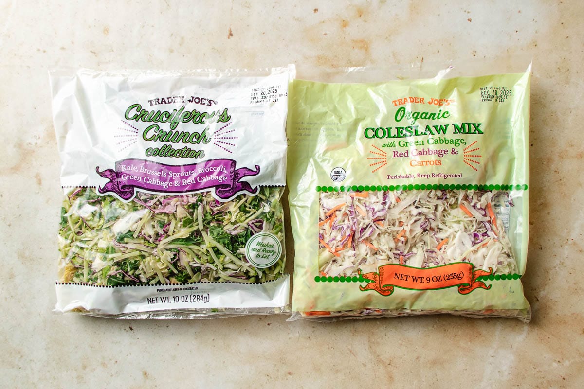 Two bags of Trader Joe’s salad mixes on a light surface: “Cruciferous Crunch Collection” and “Organic Coleslaw Mix”—perfect bases for creating a flavorful spring roll in a bowl with fresh veggies like cabbage, kale, Brussels sprouts, and carrots.
