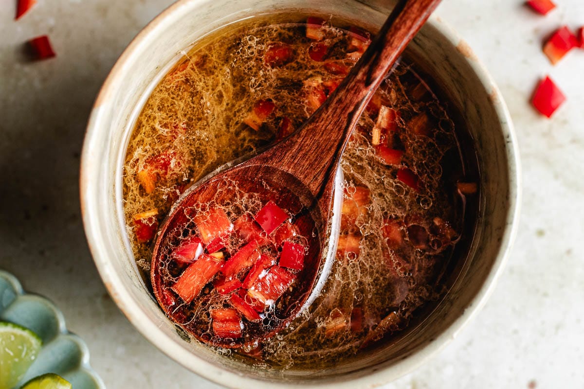 A wooden spoon holds diced red chilies above a bowl filled with Thai sweet chili lime dressing—a light brown, oily liquid with more chopped chilies. Lime wedges are partially visible on the side.