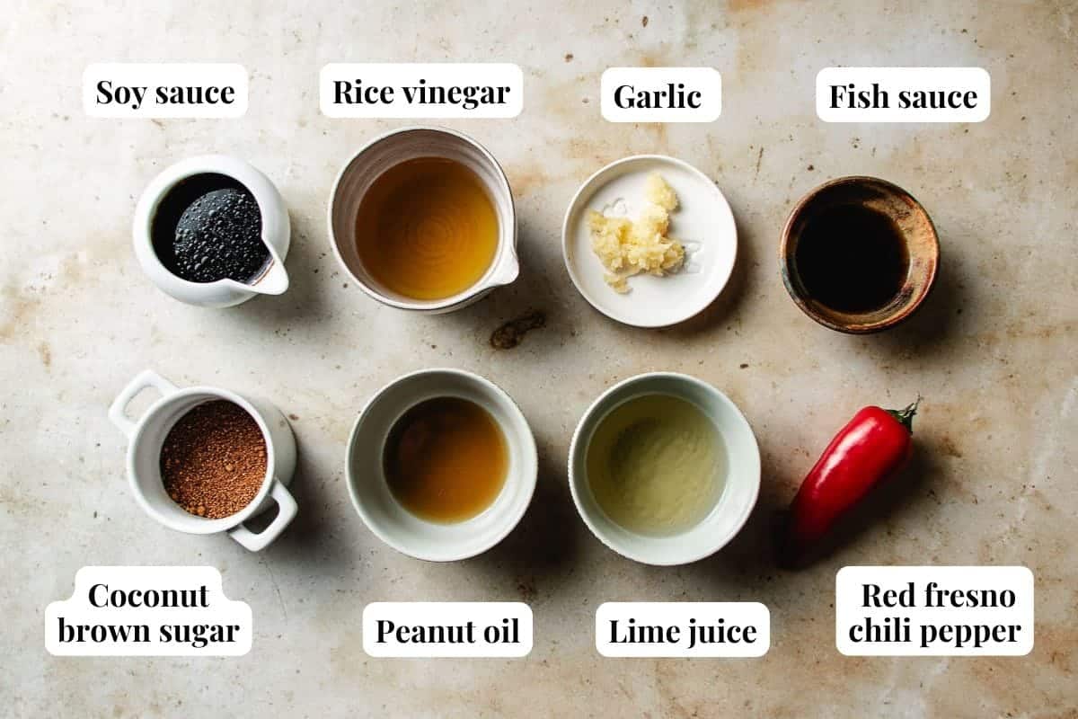 Various small bowls and containers hold soy sauce, rice vinegar, minced garlic, fish sauce, coconut brown sugar, peanut oil, lime juice, and a whole red fresno chili pepper—perfect ingredients for a Thai sweet chili lime dressing—each labeled on a light surface.