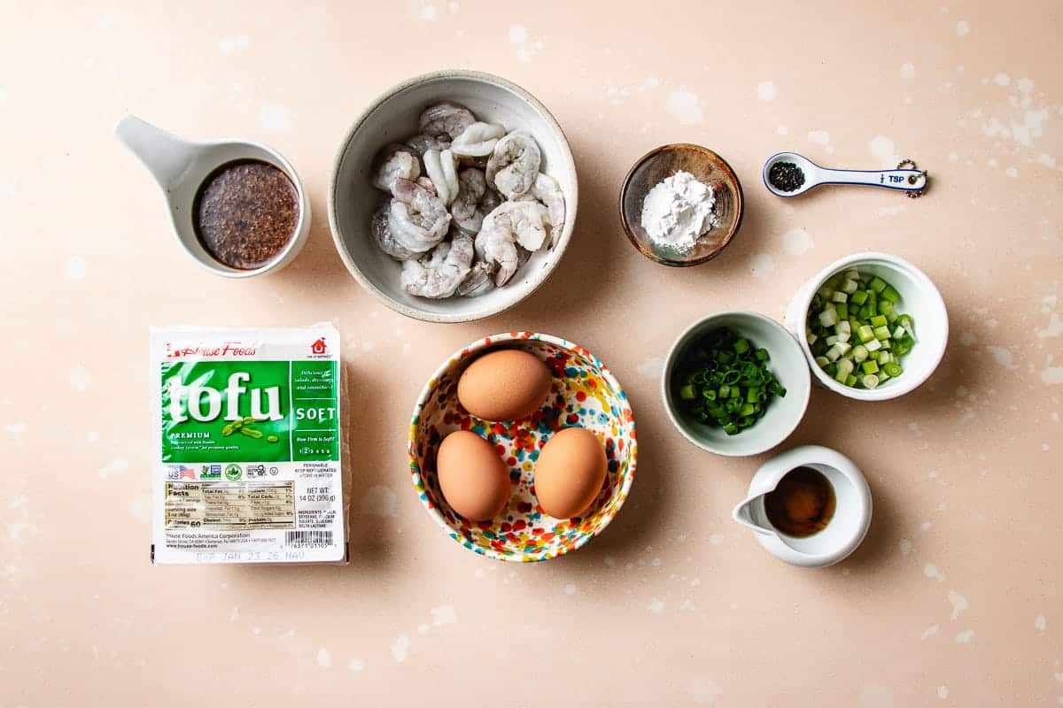 A flat lay of ingredients for a Shrimp with Tofu Bowl, featuring shrimp, soft tofu, two eggs, chopped green onions, sliced green vegetables, a bowl of starch, soy sauce, ground pepper, and oyster sauce mixture on a light countertop.