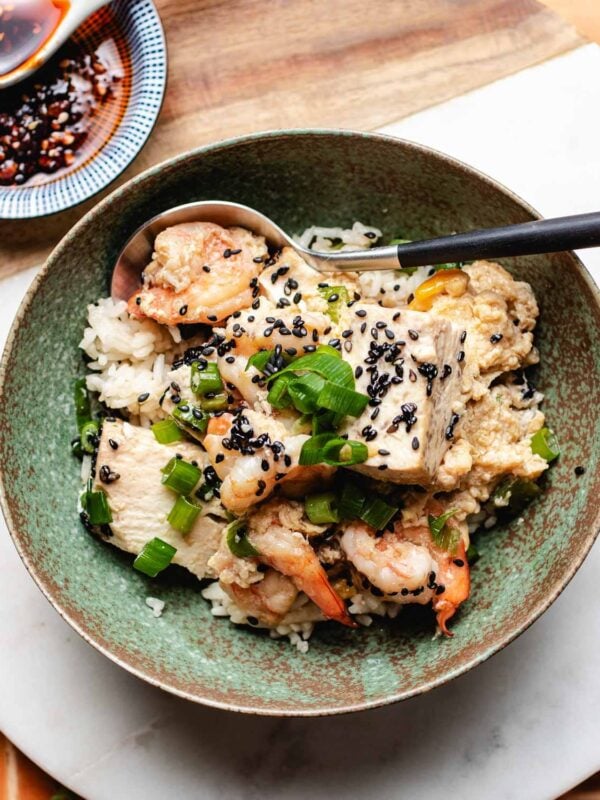 A Shrimp with Tofu Bowl features rice topped with shrimp, tofu, chopped green onions, black sesame seeds, and sauce, with a spoon resting inside. A small dish of dipping sauce is visible in the background.