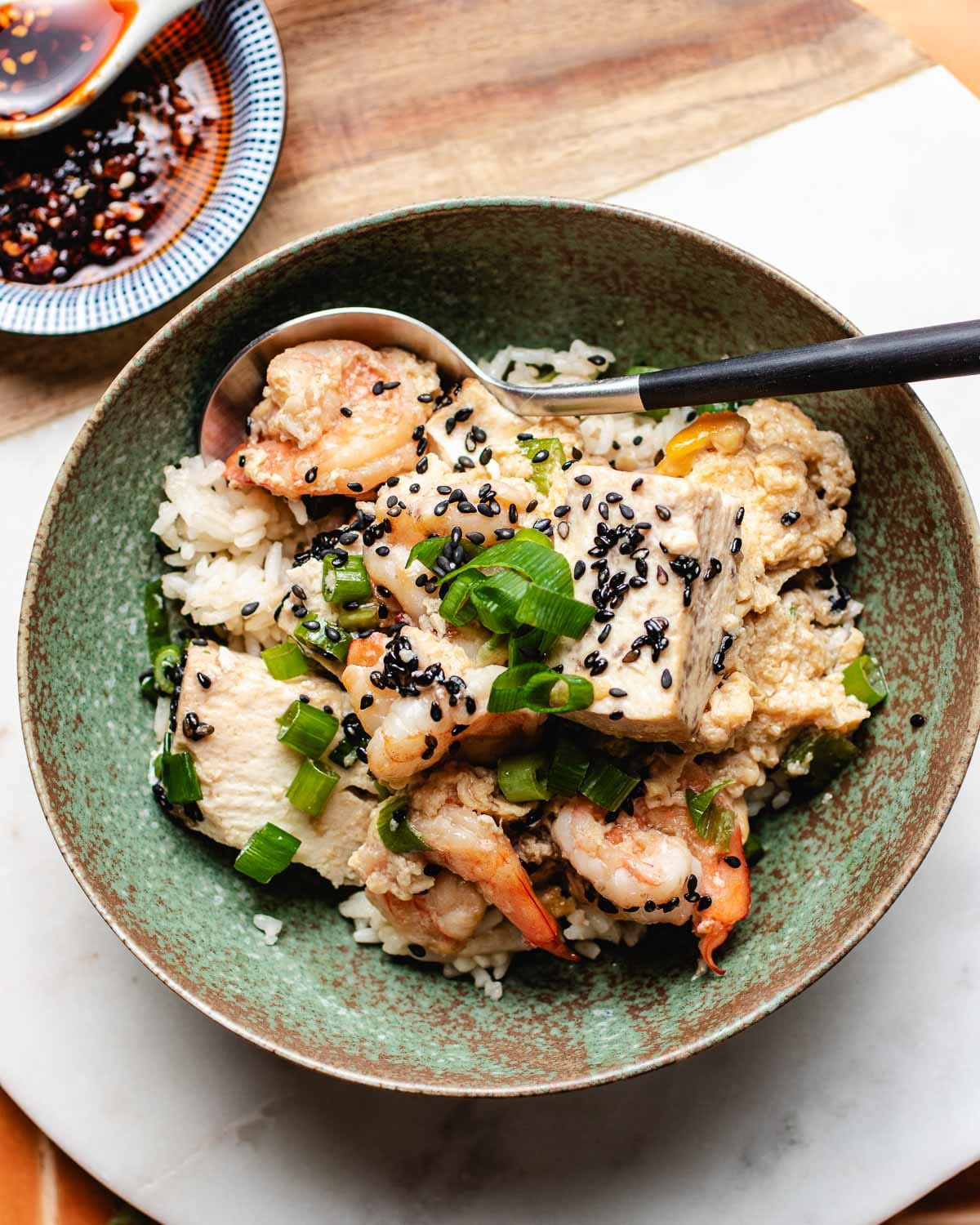 A Shrimp with Tofu Bowl features rice topped with shrimp, tofu, chopped green onions, black sesame seeds, and sauce, with a spoon resting inside. A small dish of dipping sauce is visible in the background.