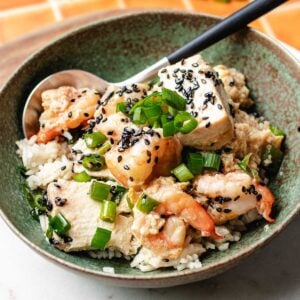 A Shrimp with Tofu Bowl featuring shrimp, tofu cubes, chopped green onions, and black sesame seed topping is served over rice, with a spoon resting inside.