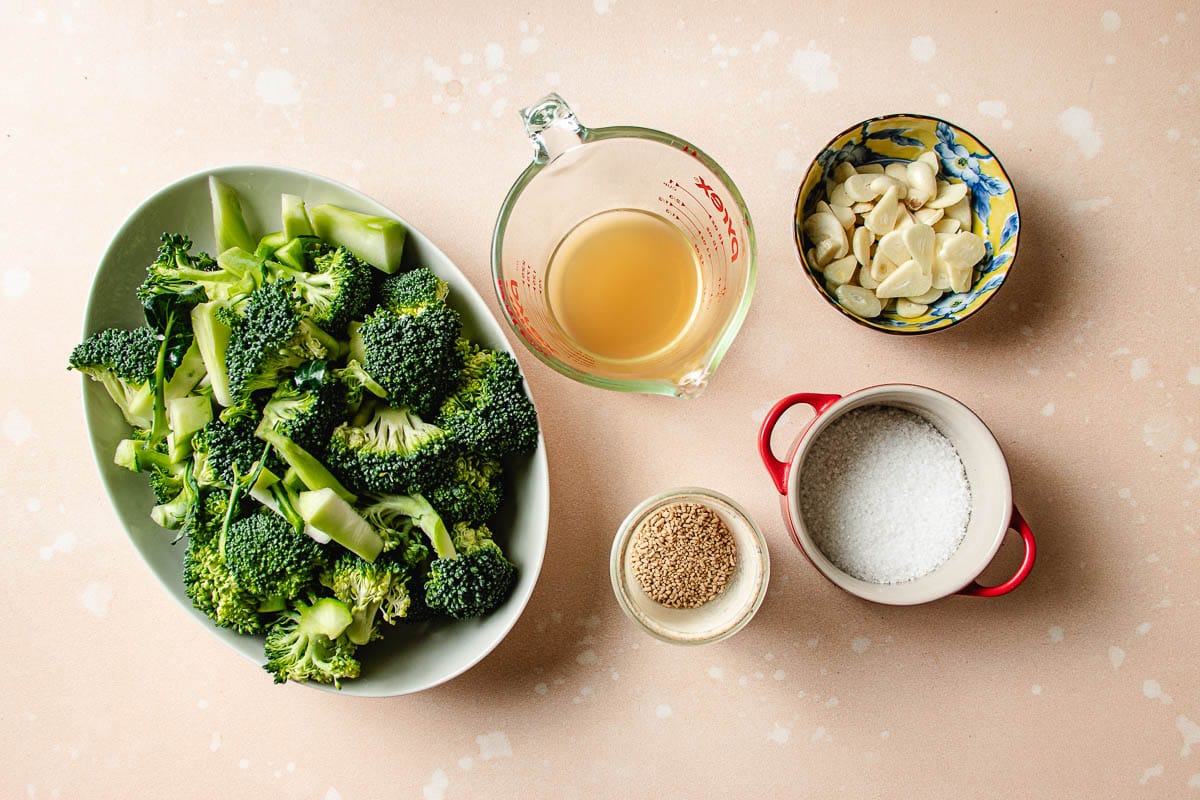 Overhead view of ingredients for Din Tai Fung Stir Fried Broccoli: broccoli florets on a plate, a measuring cup of broth, sliced garlic in a small bowl, salt, and bouillon powder on a light surface.