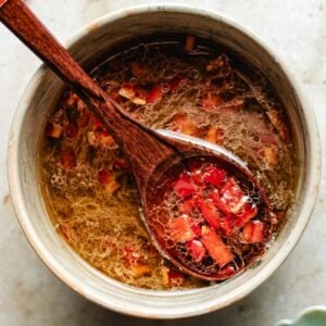 A wooden spoon holds diced red chili pepper pieces over a salad dressing bowl mixture with Thai sweet chili lime dressing in liquid, viewed from above.