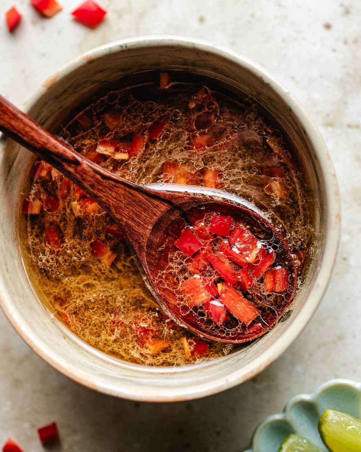 A bowl of clear, golden Thai sweet chili lime dressing with finely chopped red chili peppers, shown with a wooden spoon scooping some out. The bowl is on a light surface, with part of a green color dish with lime slices visible in the corner.