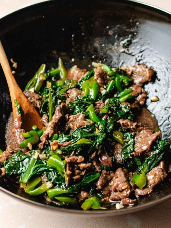 A wok filled with Beef with Chinese broccoli, green vegetables, and savory sauce. A wooden spoon rests inside, mixing the glossy, freshly cooked ingredients to perfection.