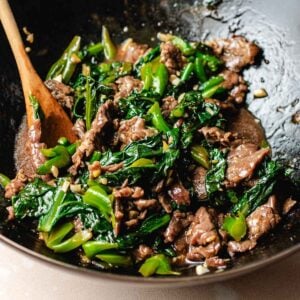 A close-up of Beef with Chinese broccoli stir-fry in a wok, featuring sliced beef, and leafy Chinese broccoli greens being mixed with a wooden spoon. The ingredients are coated in a glossy sauce.