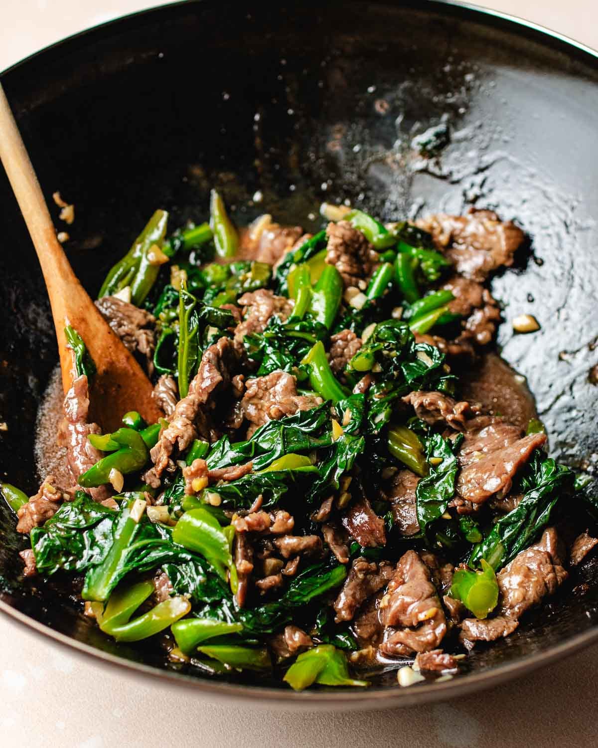 A wok filled with Beef with Chinese broccoli, green vegetables, and savory sauce. A wooden spoon rests inside, mixing the glossy, freshly cooked ingredients to perfection.