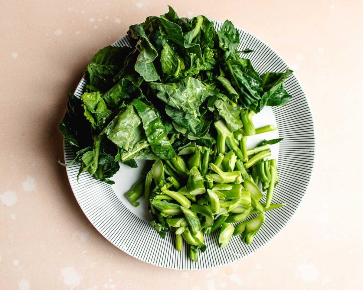 A white plate with a black striped rim holds chopped Chinese broccoli leafy greens arranged stems separately from leafy parts on a light beige surface, similar to toppings served with beef with Chinese broccoli.