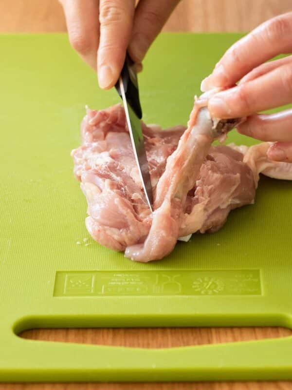 A person demonstrates how to debone chicken thighs by using a knife to cut meat from a bone on a green cutting board.