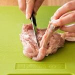 Hands demonstrating how to debone chicken thighs using a knife on a green cutting board.