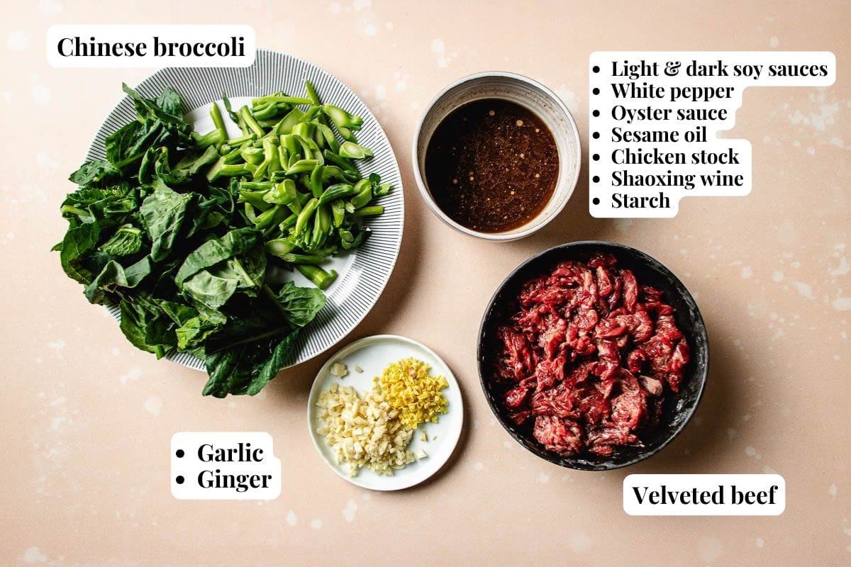 A flat lay of labeled ingredients for Beef with Chinese broccoli: chopped Chinese broccoli, minced garlic and ginger, velvety beef slices, and a bowl of mixed sauces and seasonings on a light background.
