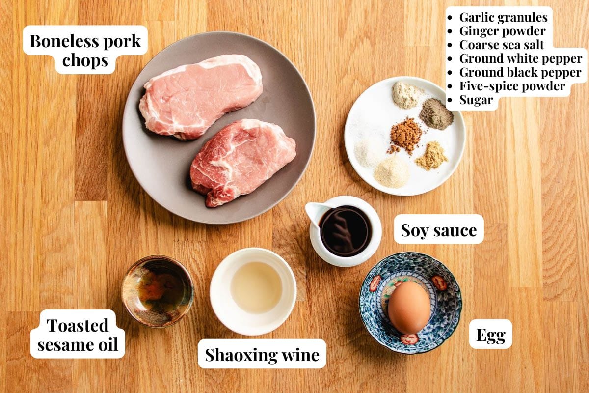 Overhead view of a plate with two raw boneless Taiwanese pork chops and five bowls containing toasted sesame oil, Shaoxing wine, soy sauce, an egg, and various spices, all labeled on a wooden surface.