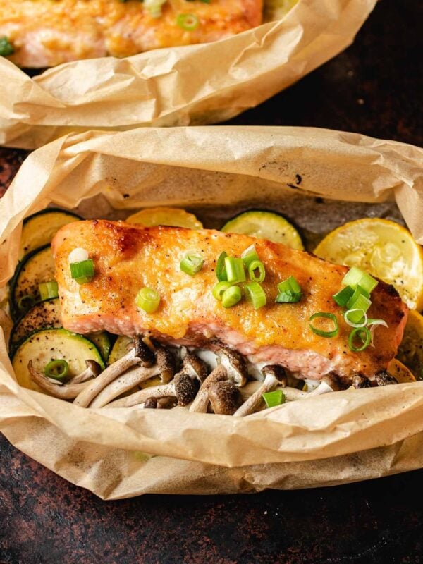A miso butter salmon fillet topped with chopped green onions sits on a bed of sliced zucchini, yellow squash, and mushrooms, all wrapped in parchment paper.