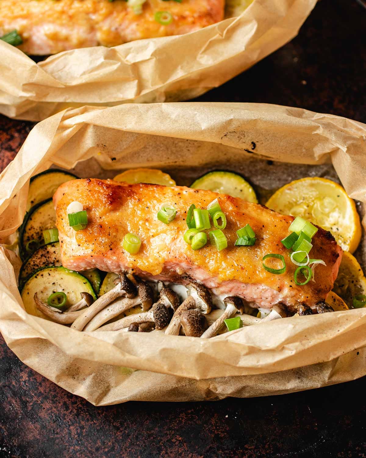 A miso butter salmon fillet topped with chopped green onions sits on a bed of sliced zucchini, yellow squash, and mushrooms, all wrapped in parchment paper.