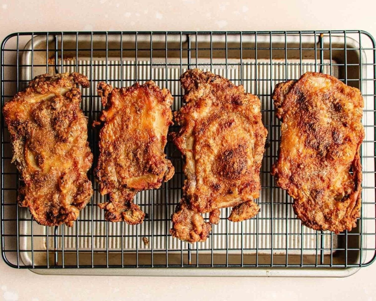Four pieces of golden-brown, perfectly crisp boneless, skin-on chicken thighs rest on a wire rack placed over a baking sheet. Perfect for those learning how to debone chicken thighs, this dish appears freshly cooked and deliciously crunchy.