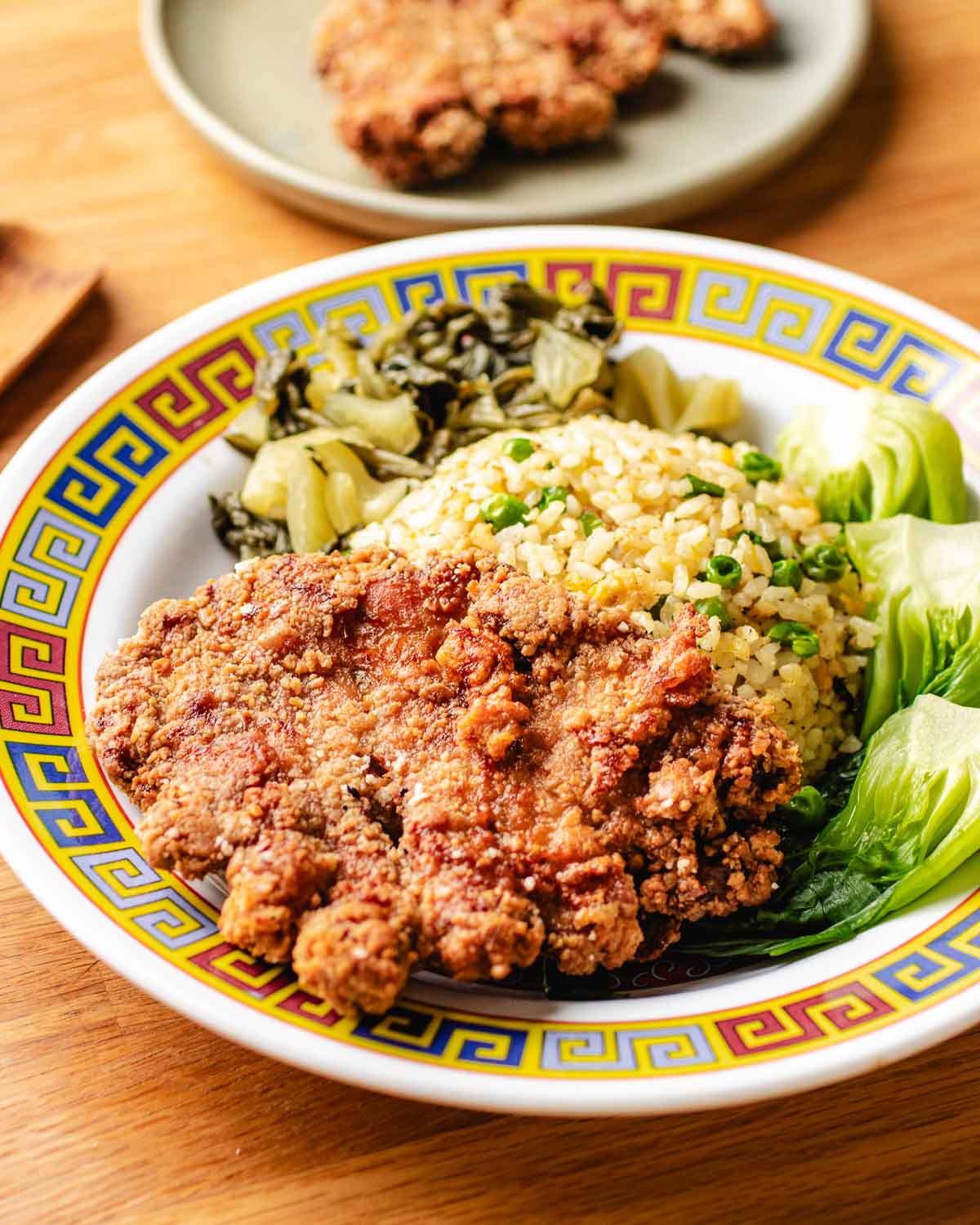 A plate with crispy Taiwanese pork chop, fried rice topped with green onions, pickled greens, and bok choy, served in a decorative bowl on a wooden table. Another pork chop is on a plate in the background.