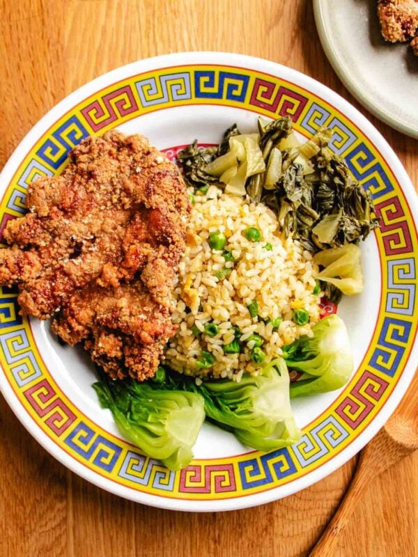 A colorful plate with crispy Taiwanese pork chop, fried rice with pickled mustard greens and blanched baby bok choy, on a patterned dish atop a wooden table.