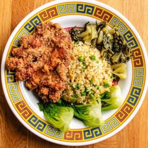 A colorful plate holds crispy Taiwanese pork chop, fried rice with peas, pickled mustard greens, and steamed bok choy, all arranged neatly on a decorative round dish atop a wooden surface.