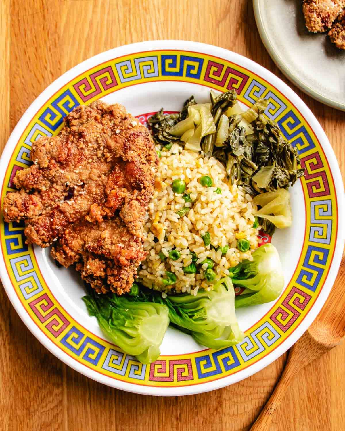 A colorful plate with crispy Taiwanese pork chop, fried rice with pickled mustard greens and blanched baby bok choy, on a patterned dish atop a wooden table.