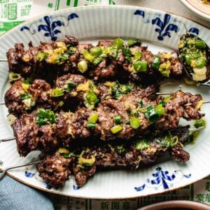 Beef skewers with ginger scallion sauce, garnished with ginger scallion sauce and a yellow sauce, served on a decorative white and blue oval plate.
