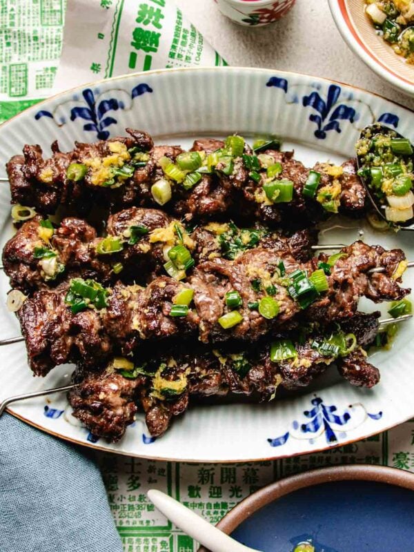 Beef skewers with ginger scallion sauce are garnished with scallion sauce and served on a white oval plate with blue designs. A spoon of topping rests on the plate, set atop Asian-themed paper.