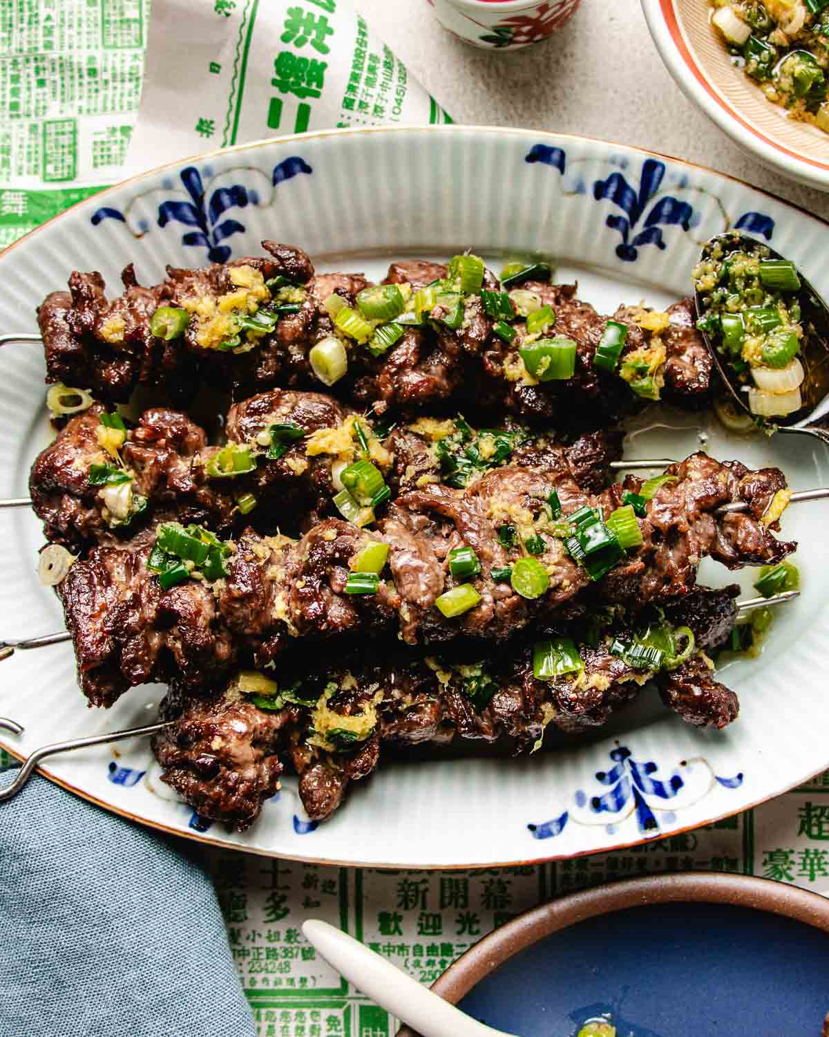 Beef skewers with ginger scallion sauce are garnished with scallion sauce and served on a white oval plate with blue designs. A spoon of topping rests on the plate, set atop Asian-themed paper.