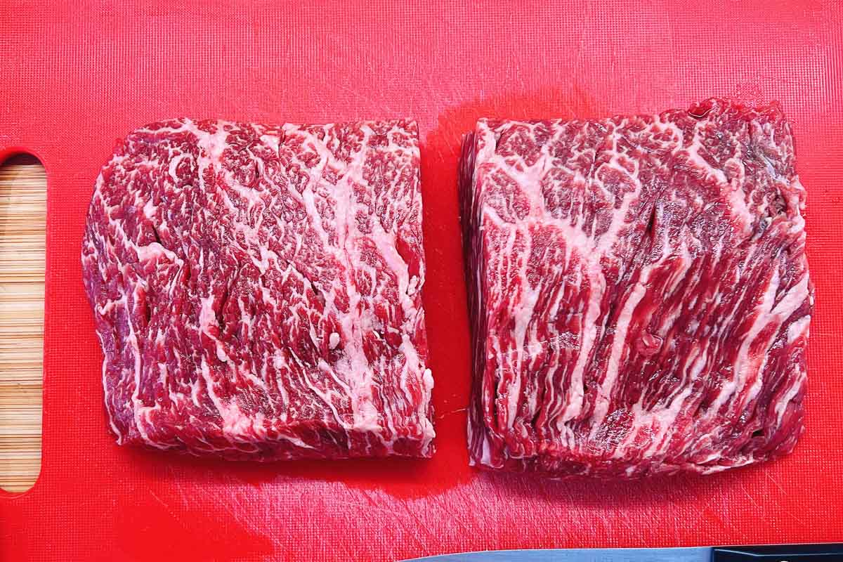 Two pieces of marbled raw beef (sirloin flat steak), perfect for beef skewers with ginger scallion sauce, are placed side by side on a bright red cutting board, with visible white fat streaks running through the meat.
