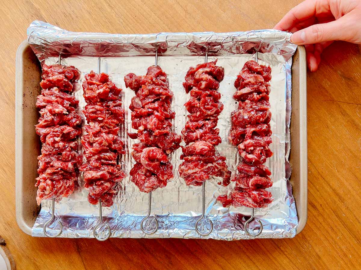 Five metal skewers threaded with raw marinated beef for beef skewers are arranged on a foil-lined baking sheet. A hand is holding the tray on the right side, all placed on a wooden surface.