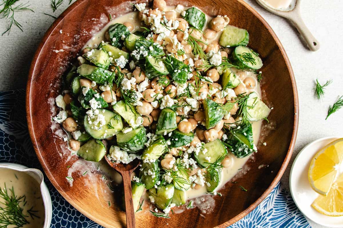 A wooden bowl filled with cucumber chickpea salad—chopped cucumbers, chickpeas, crumbled blue cheese, fresh dill, and creamy dressing. A wooden spoon, lemon wedges, and extra dressing are nearby.