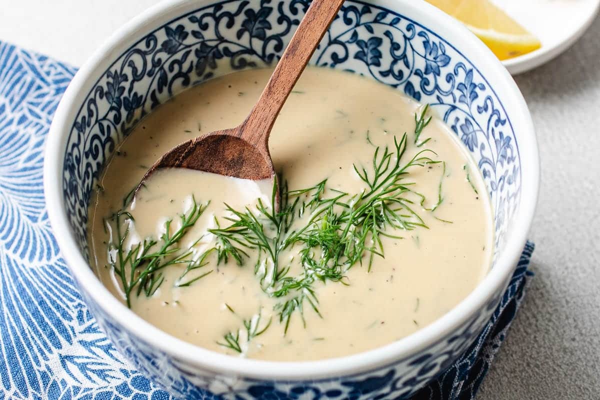 A decorative blue and white bowl filled with creamy Tahini dill dressing, garnished with fresh dill and stirred with a wooden spoon; a lemon wedge is visible in the background.