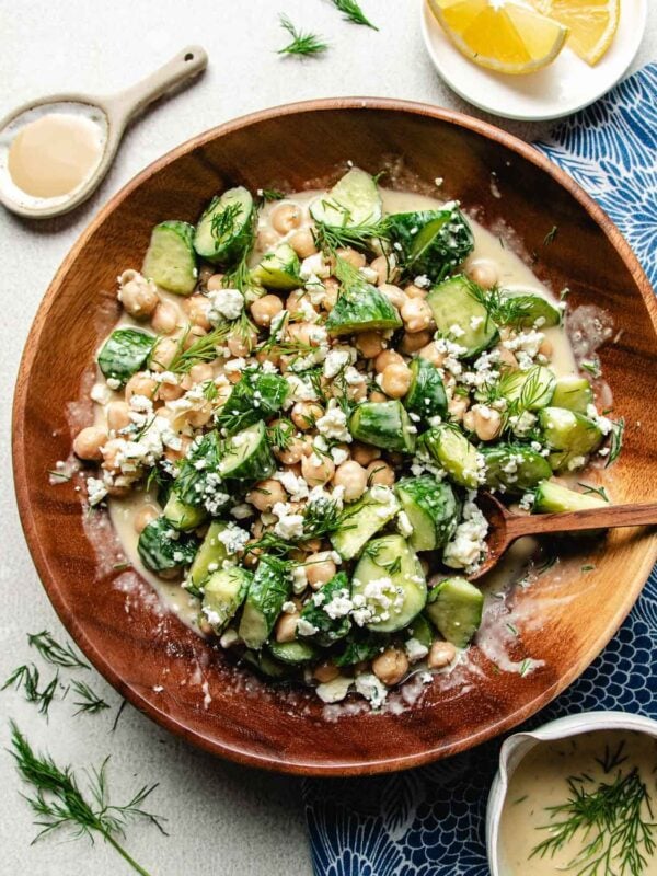 A wooden bowl of cucumber chickpea salad with fresh dill, and crumbled blue cheese sits with a wooden spoon. Nearby are a small bowl of creamy dressing and lemon wedges on a blue patterned cloth.