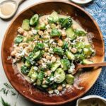 A wooden bowl filled with cucumber chickpea salad—sliced cucumbers, chickpeas, crumbled blue cheese, fresh dill, and creamy tahini dressing. A wooden spoon rests in the bowl with fresh dill scattered around.