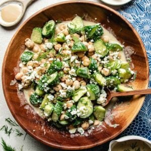 A wooden bowl filled with cucumber chickpea salad—sliced cucumbers, chickpeas, crumbled blue cheese, fresh dill, and creamy tahini dressing. A wooden spoon rests in the bowl with fresh dill scattered around.
